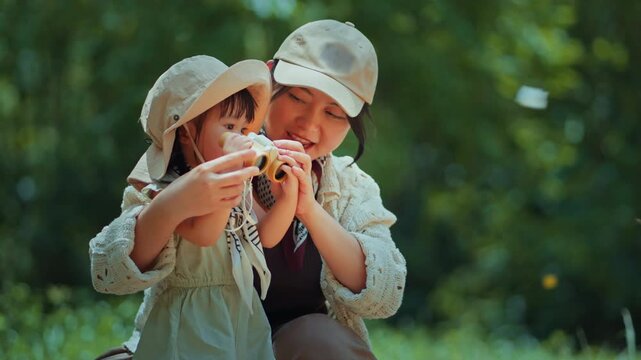 Asian mother and toddler daughter with binoculars exploring a lush forest together to see butterflies. This heartwarming nature adventure captures discovery, family bonding, and education