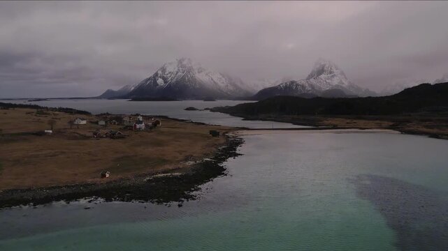 Serene coastal village nestled beneath snow-capped mountains under cloudy skies