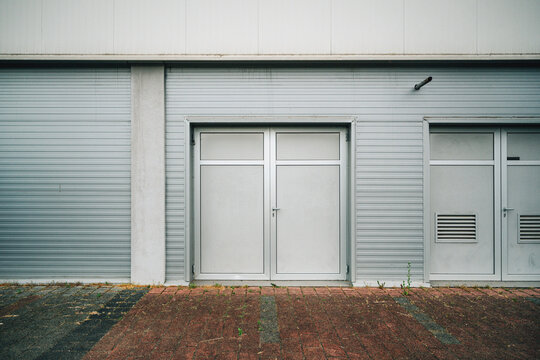 Minimal industrial scene featuring metal double doors, roller shutters and a service door on a pale warehouse facade, set against weathered brick pavement.