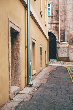 A quiet narrow European alley framed by a weathered yellow building, blue downspout and arched black doorway. Textured stucco walls, cobblestone pavement and muted tones.
