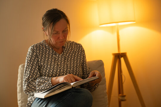 Middle-aged woman reading book on sofa at home, calm evening atmosphere and thoughtful lifestyle moment