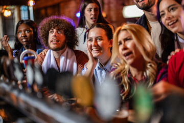 Fototapeta premium Cheerful young fans looking at a screen and celebrating a soccer match victory in a pub.