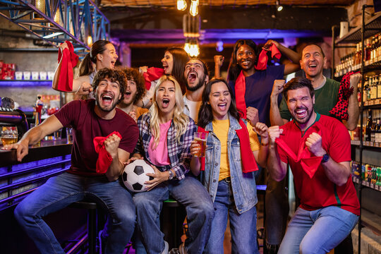 Ecstatic sports fans with red scarves and jerseys jumping and celebrating a soccer goal in a pub.