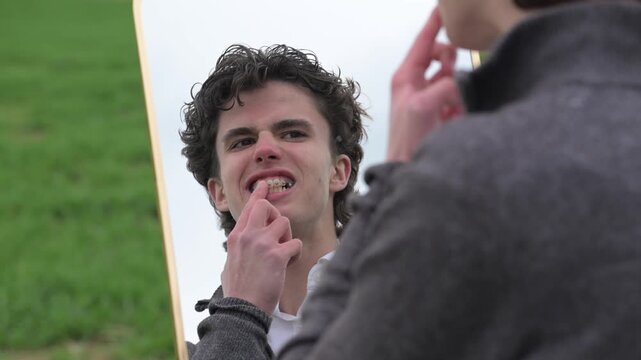 Young man examining his teeth in mirror and touching them with finger outdoors in natural light. Close-up dental care concept with healthy lifestyle atmosphere.
