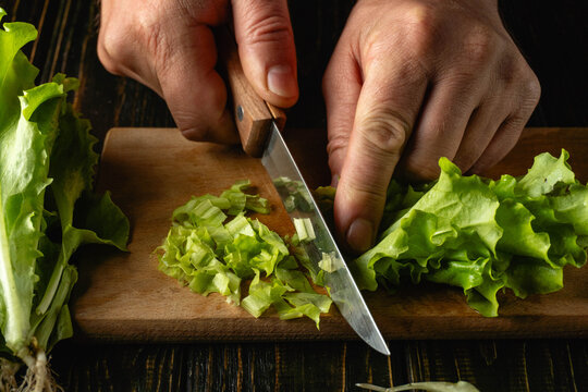 Two hands cut fresh lettuce on a wooden board. The green lettuce is neatly placed while being prepared for salad. The kitchen setting shows preparation for cooking or meal