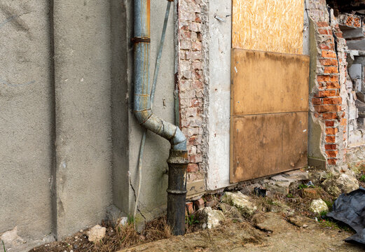 Old metal drainpipe attached to a cracked plaster and exposed brick wall beside a boarded doorway and crumbling masonry, highlighting architectural decay, abandoned building detail, and urban deterior