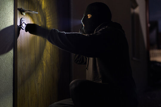 Vandalism concept. Unrecognizable thief in black clothes trying to unlock door with picklock, using flashlight at nighttime