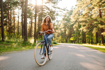 Obraz premium A woman rides a blue bicycle on a path through a green forest. She smiles as she pedals under the trees on a sunny day. The scene shows a moment of active fun and joy.