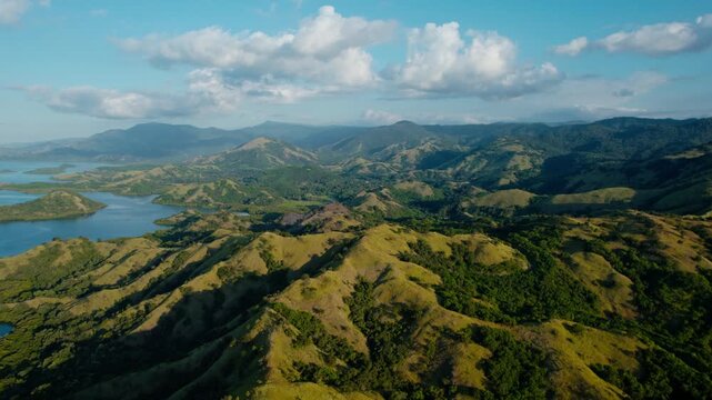High angle drone shot flying forward over green savanna hills overlooking calm blue sea bays in Riung 17 Islands National Park, Flores.
