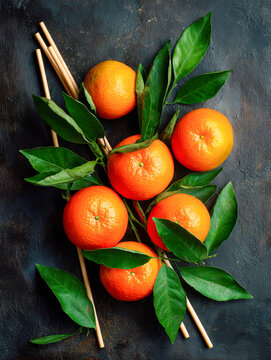 Bright ripe mandarins with fresh green leaves arranged on rustic dark textured surface with natural wooden sticks in a vibrant colorful composition