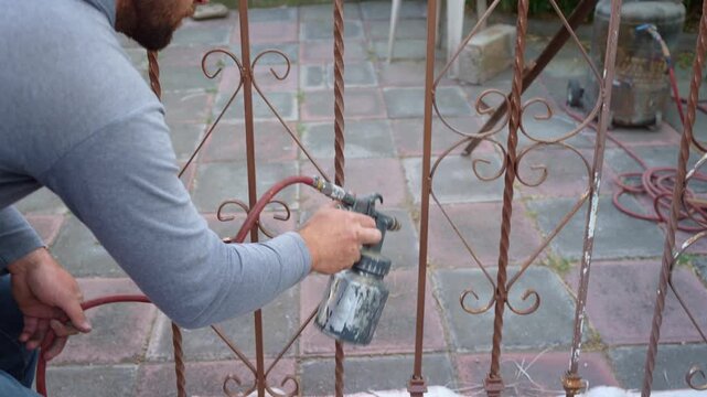 Man using a paint sprayer to coat metal window security bars, restoring exterior protection at home. Home maintenance, renovation work, DIY improvement