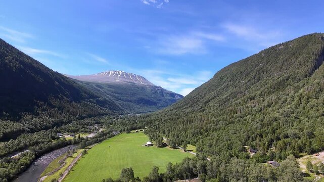 Aerial slow motion drone footage of mountain river flowing through forested valley toward Gaustatoppen peak in Telemark, Norway. Summer landscape with clear blue sky and lush green vegetation.