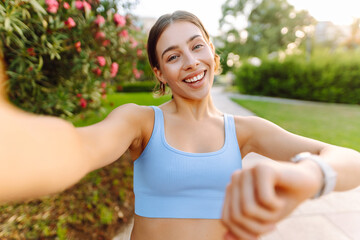 Fototapeta premium Portrait of a smiling woman in sportswear taking a selfie in a sunny park. A young athlete enjoys relaxing after a workout. Concept for sports and blogging. Active lifestyle.