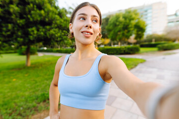 Fototapeta premium Portrait of a smiling woman in sportswear taking a selfie in a sunny park. A young athlete enjoys relaxing after a workout. Concept for sports and blogging. Active lifestyle.