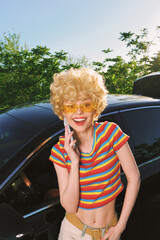 Young woman with curly blonde hair stands beside a dark car, wearing a colorful striped crop top...