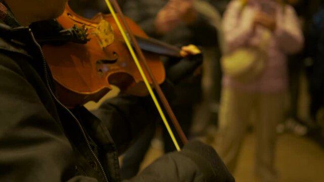 Street musician playing violin at night. Close-up of a talented violinist playing a classical instrument on a city street at night, with a blurred crowd of people in the background