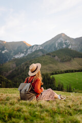 Naklejka premium A smiling woman with a backpack and hat stands on a green hill at sunset. The young woman feels free, enjoying the mountain views on a sunny day. Concept of travel and adventure.
