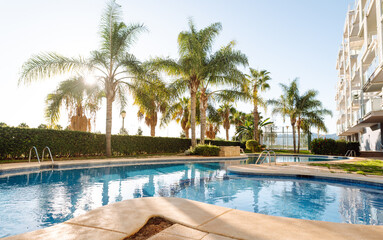 Fototapeta premium The setting shows a swimming pool with steps leading into the water. Surrounding the pool are green plants and trees. The sun shines brightly, creating reflections on the water surface.