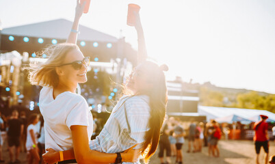 Two friends are having fun and holding drinks at a music festival. Young women are enjoying the festival on the beach, dancing at sunset with drinks. Concept of vacation, relaxation, and fun.