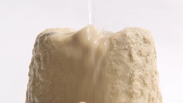Thin water stream falls from above onto beige granular mound, gradually eroding the peak and creating flowing channels as the sandy material dissolves and collapses inward.