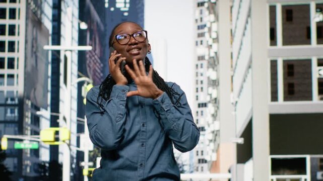 Smiling black woman engaged in phone call on mobile device, strolling through bustling cityscape. Outdoor urban scene captures fashionable pedestrian enjoying casual lifestyle while chatting.