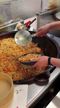 A cook serves fresh Uzbek pilaf from a large cauldron onto a plate.