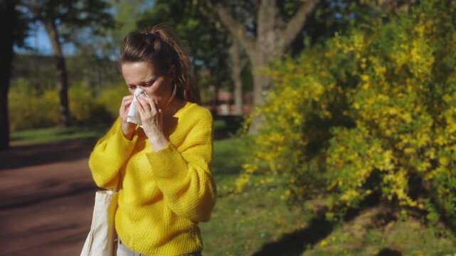 A woman in a yellow sweater deals with hay fever symptoms, blowing her nose with a tissue while walking through a sunlit park with blooming yellow bushes.