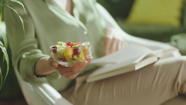 A woman relaxes in a white chair, reading a book while enjoying a healthy fruit salad, capturing a moment of mindful eating and peaceful leisure.