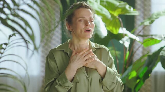 Close-up of a calm woman in a green shirt with her hands on her chest, focusing on her breath to find inner peace and emotional balance in a peaceful indoor setting.