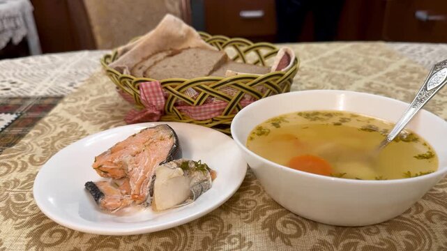 Fish soup with trout and bread basket on patterned tablecloth, clear herb broth with carrot slice, seared trout fillet on white