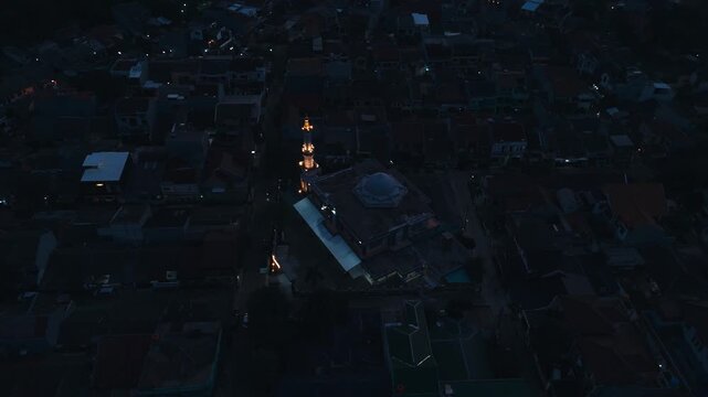 Aerial top view of a grand mosque illuminated at night surrounded by dark residential village houses in evening dawn