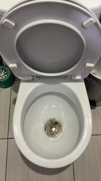 White porcelain toilet bowl stained with urine, overhead view in tiled bathroom showing toilet brush, gray plumbing pipe, used