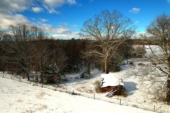 defaultAerial drone view of snow-covered rural Virginia farmland with collapsed barn, bare winter trees, wooden fence, and dramatic blue sky in Pittsylvania County