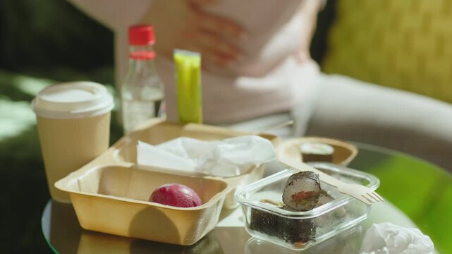 A woman rubs her stomach in physical distress behind a table cluttered with sushi takeout containers, highlighting the consequences of unhealthy takeaway food.