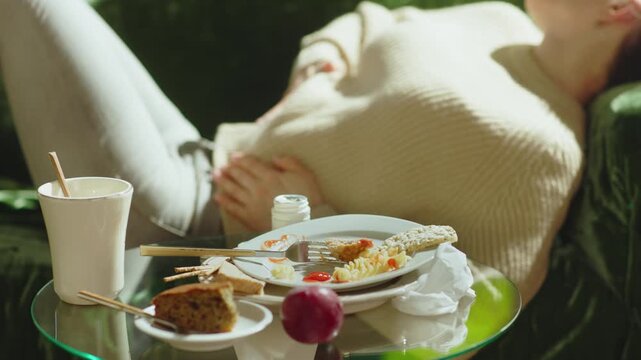 A woman lies on a green couch in physical discomfort, holding her stomach next to a table of messy food scraps and medicine, illustrating the effects of a poor diet.