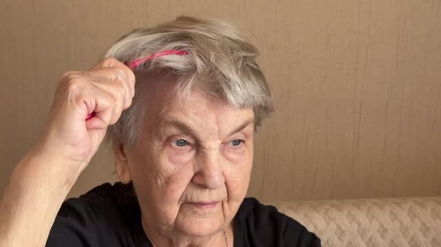 Elderly grandmother using red massager close-up, seated on sofa, grey hair with light sparkle, gentle scalp kneading