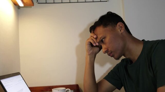 Exhausted Young Asian professional feeling burnt out and confused while staring at a laptop screen in a cozy workspace.