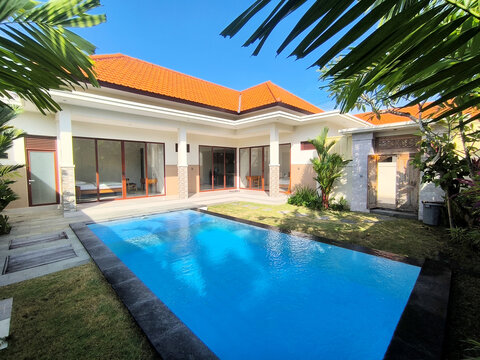 Beautiful private pool in a modern Balinese villa with orange tile roof, surrounded by lush tropical gardens and traditional carved wooden doors, under bright blue skies.