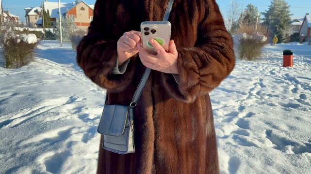 Woman in mink coat texting closeup, hands holding phone in snowy park, severe frost on fur, crossbody bag at side
