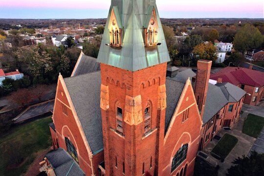 Aerial drone view of First Baptist Church's illuminated historic red brick steeple with copper patina roof glowing at dusk over Danville Virginia