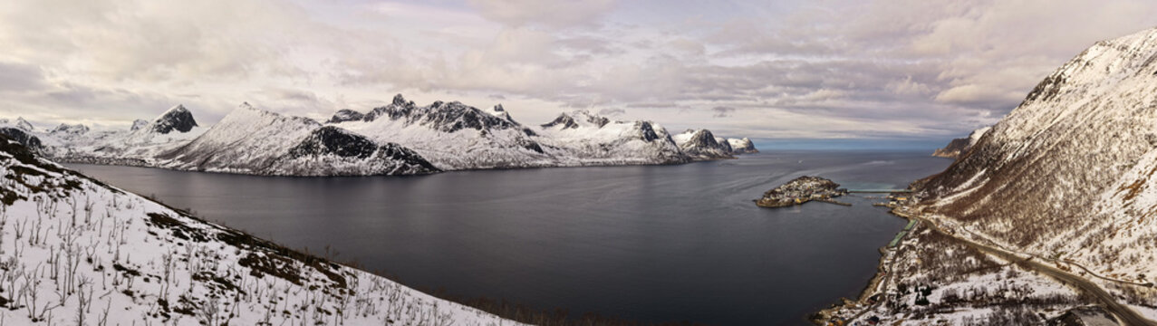 Drones fly over the northern region of Norway inn Husoy, revealing stunning views of fjords surrounded by snow-covered mountains and a calm sea under a cloudy sky.