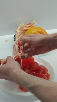 Close-up view of male hands peeling a vibrant red grapefruit, removing the white pith and skin to reveal the tender, juicy fruit segments, ready for a nutritious and vitamin-rich breakfast or snack on