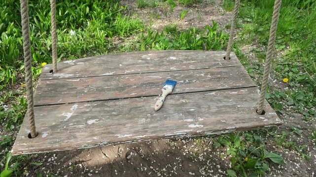 A rustic and worn wooden swing with peeling paint, hanging by sturdy ropes, subtly moves in an outdoor setting with vibrant green grass, showcasing an abandoned paint brush with blue traces on its