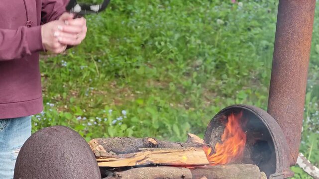 Detailed view of a person's hands diligently stoking a robust wood fire in a rustic, homemade outdoor barbecue setup, enhancing the burning logs and rising smoke with a metal spatula, reflecting the
