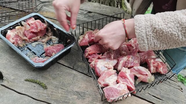 A person is carefully placing seasoned chunks of raw pork or beef from a plastic tray onto a metal BBQ grilling rack, set on an old wooden surface outdoors, getting ready for cooking on an open fire