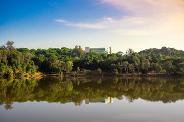 Fototapeta premium Scenic lake reflecting dense lush green trees and modern building in distance