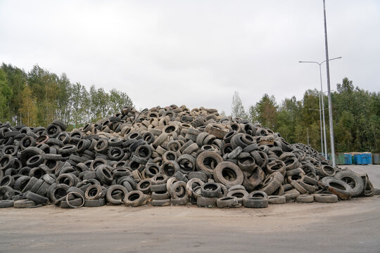 Many discarded rubber tires form a huge mound on a paved lot at an industrial waste facility. Trees line the background under an overcast daytime sky.