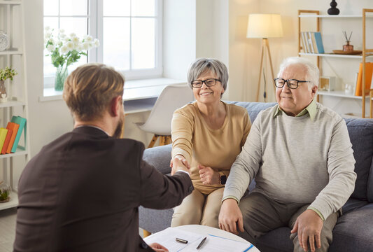 Senior couple discussing retirement plan or bank savings with professional pension agent or financial adviser. Happy healthy mature husband and wife shake hands with consultant sitting on sofa at home