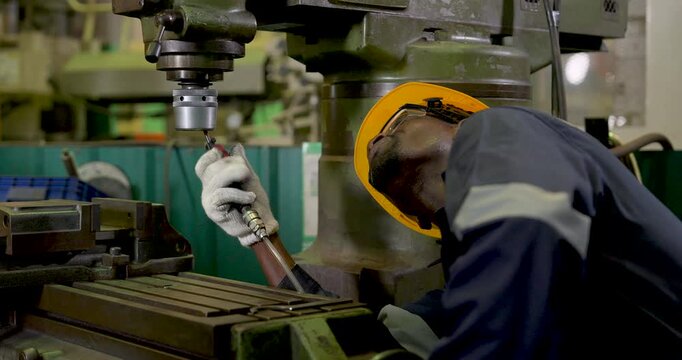 African adult male technician leans under milling machine spindle, using air blow gun for maintenance cleaning and inspection during safe equipment setup in workshop slow motion sequence