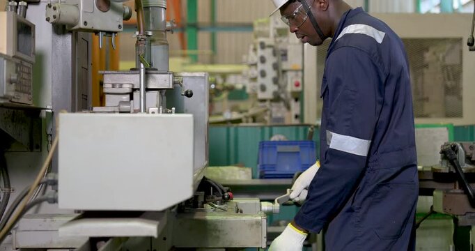African adult male operator guides machine table during metalworking process in workshop, monitoring industrial equipment movement with glove for safe precise production control.